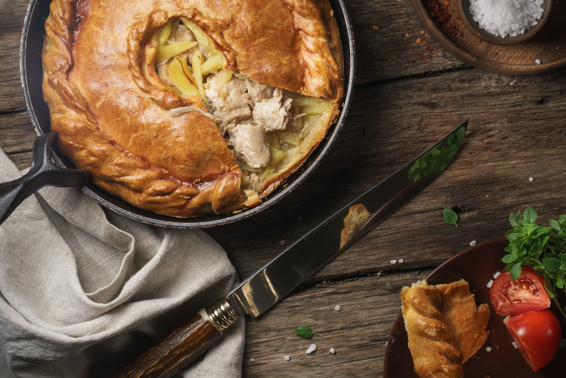 A golden chicken and leek pie in a cast iron skillet with flaky pastry crust, served alongside fresh herbs and tomatoes on a rustic wooden table.