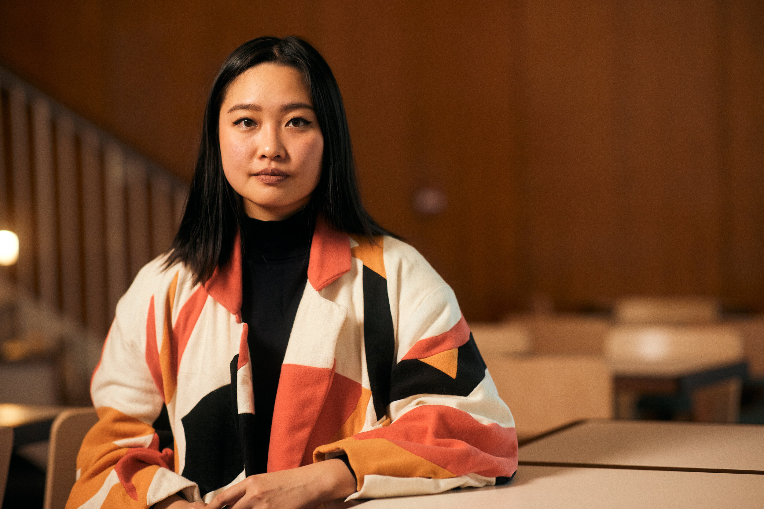 An Asian woman in a colorful geometric jacket sits at a restaurant table, looking directly at the camera with a serious expression.