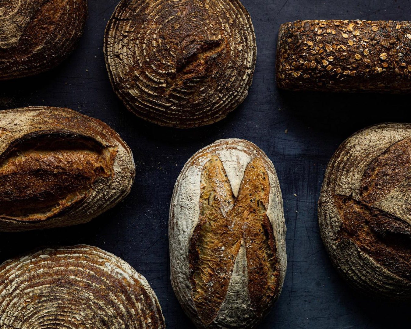 Various artisanal sourdough loaves with decorative scoring patterns arranged on a dark wooden surface.