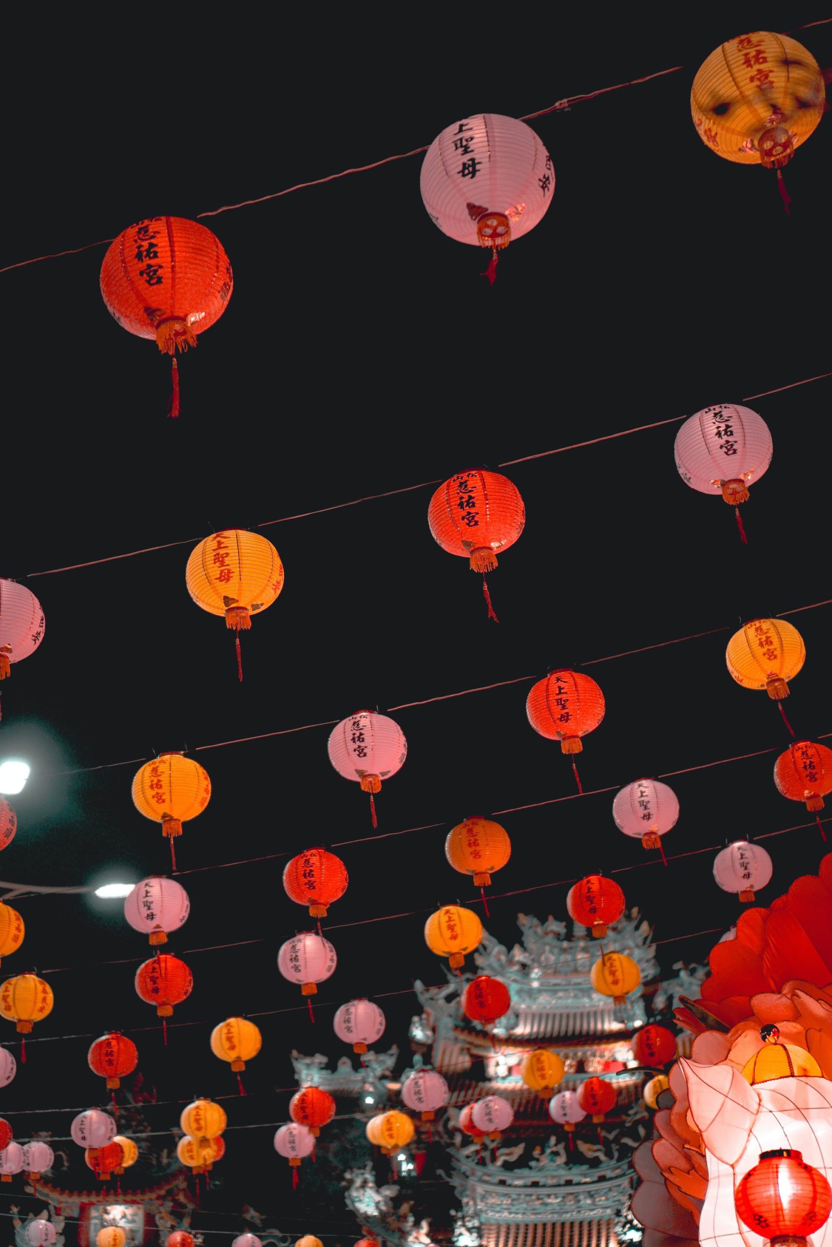 Colorful red, pink, and orange Chinese lanterns with calligraphy hang from strings against a dark night sky above traditional temple rooftops.