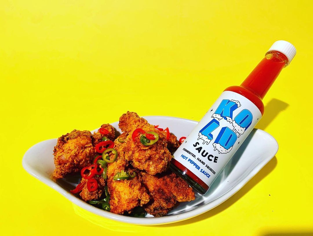 A plate of golden fried chicken topped with sliced chilies sits beside a bottle of Kold Sauce hot pepper sauce on a bright yellow background.
