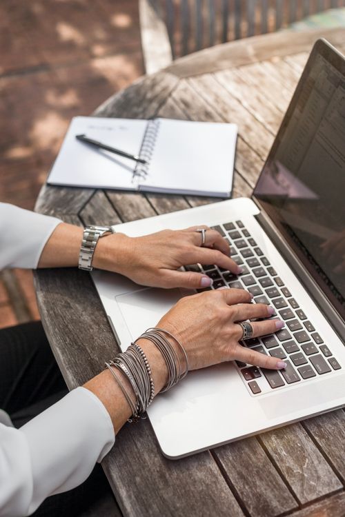 Hands typing on a laptop keyboard at an outdoor wooden table with a notebook and pen nearby.