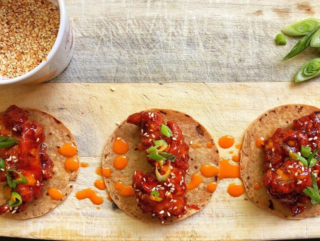 Three corn tortillas topped with red sauce, orange dots, sesame seeds, and green herbs on a wooden cutting board.