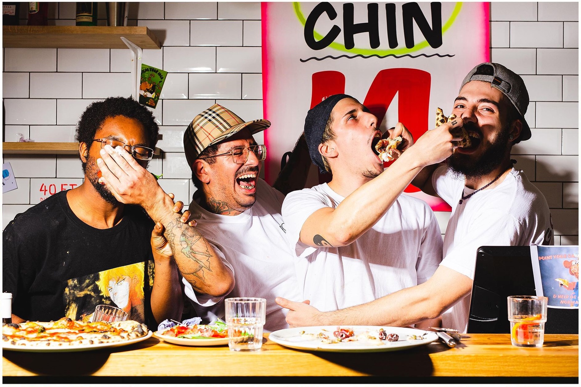 Four friends laughing and enjoying pizza together at a restaurant table with white subway tile walls in the background.