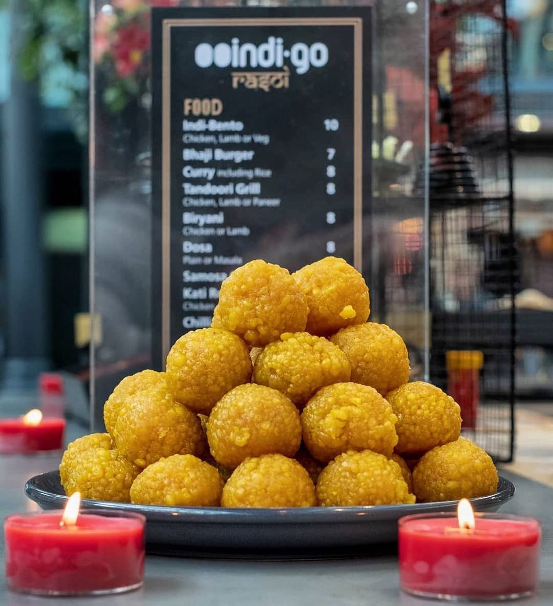 Golden laddoo sweets arranged on a dark plate with lit red candles beside a restaurant menu board.