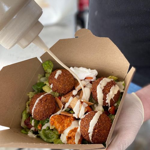 A takeout box filled with falafel balls, fresh greens, and white sauce being held by someone's hand.
