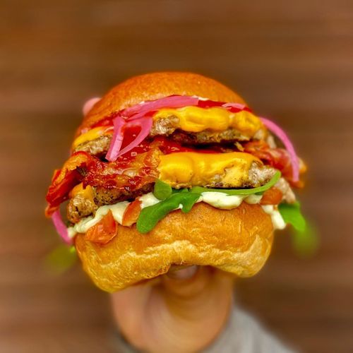 A hand holds a towering burger with multiple beef patties, melted cheese, pickled onions, and fresh greens on a golden bun.