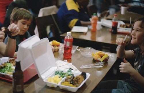 Two children eating packed meals with vegetables and drinks at a school cafeteria table.