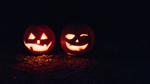 Two glowing jack-o'-lanterns with carved faces sit on fallen autumn leaves in the dark, creating an eerie Halloween atmosphere.