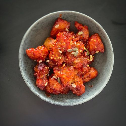 A speckled ceramic bowl filled with glossy, red-orange gochujang-glazed cauliflower topped with sesame seeds and chili flakes.