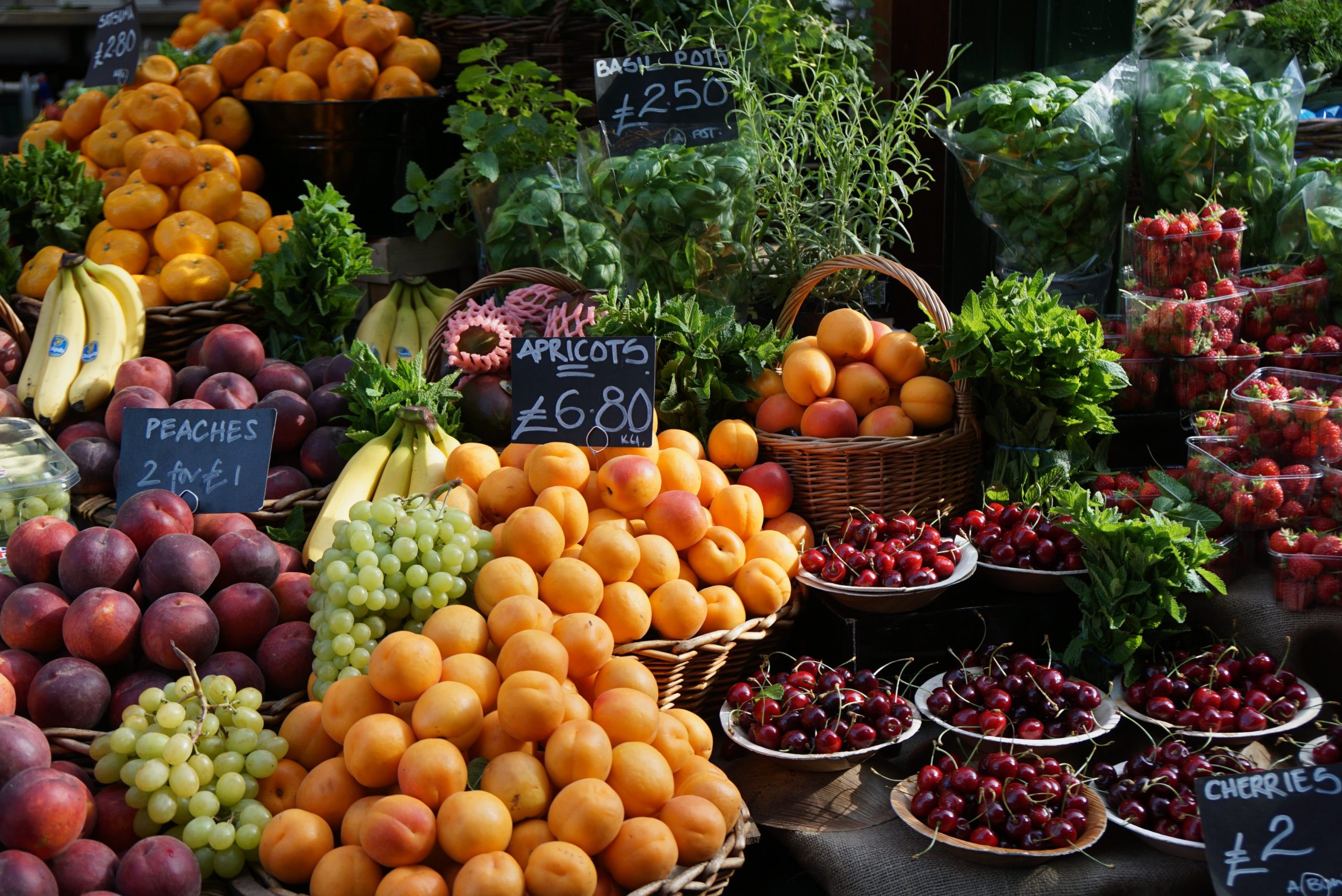 A vibrant farmers market display featuring fresh oranges, peaches, apricots, cherries, grapes, and leafy greens with handwritten price signs.