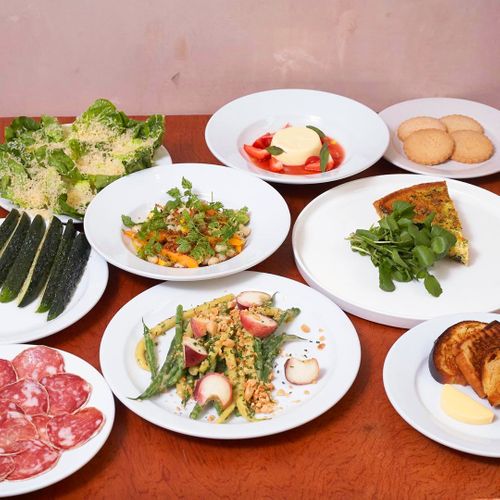 Multiple white plates displaying gourmet meal components including salad, vegetables, meat, and dessert arranged on a wooden table.
