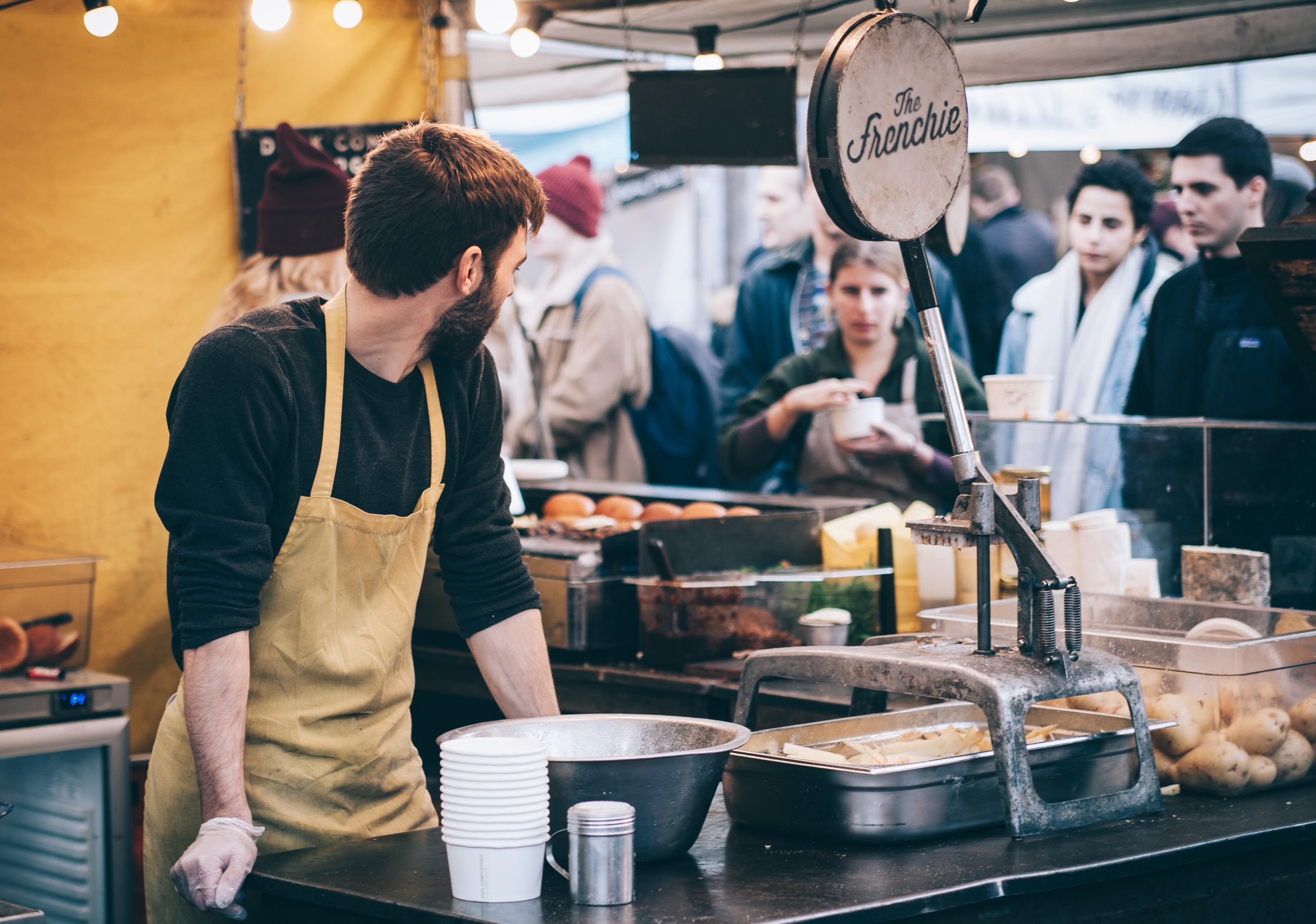 A chef in an apron prepares food at "The Frenchie" food stall while customers wait in line at a bustling market.