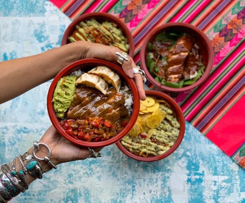 Hands holding colorful bowls of Mexican food including guacamole, salsa, and other traditional dishes on a vibrant striped tablecloth.