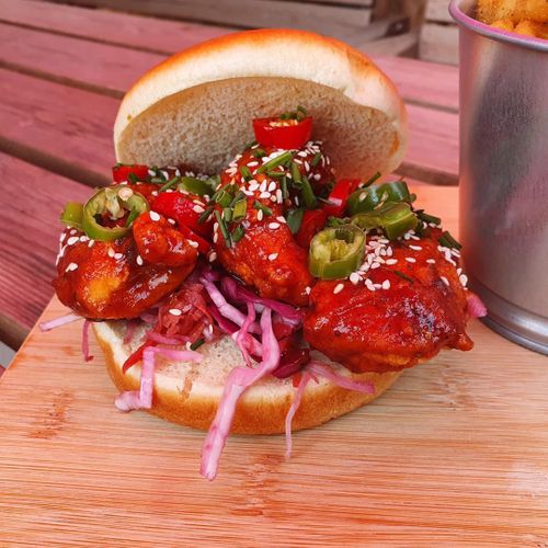 A loaded sandwich with glazed meat, pickled onions, jalapeños, and sesame seeds on a brioche bun, served on a wooden cutting board.