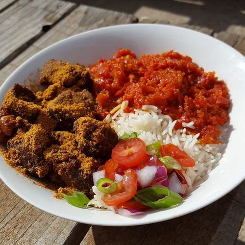 A white bowl filled with spiced fried chicken, tomato-based stew, white rice, and fresh diced vegetables on a wooden table.