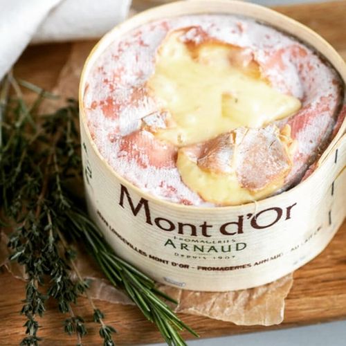 A wheel of creamy Mont d'Or cheese with melted center sits on a wooden board, garnished with fresh rosemary sprigs.