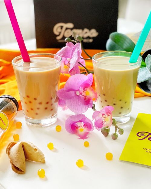 Two bubble tea drinks with tapioca pearls and colorful straws sit beside pink orchid flowers and fortune cookies on a white table.