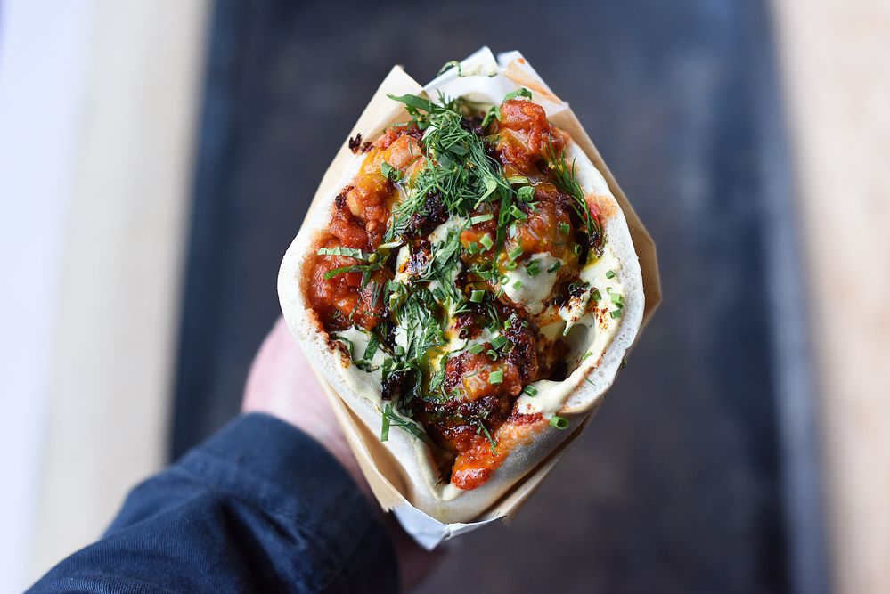 A hand holds a soft pita sandwich filled with grilled meat, fresh herbs, tomatoes, and white sauce from Shuk restaurant.
