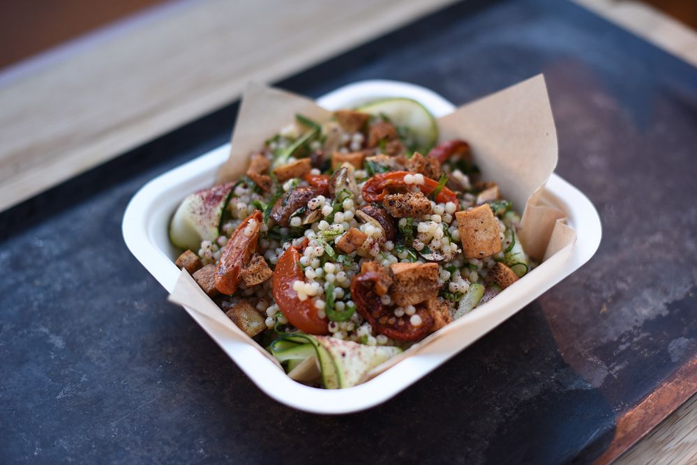 A colorful Middle Eastern grain salad with bulgur, dried tomatoes, herbs, and croutons served in a white container with cucumber slices.