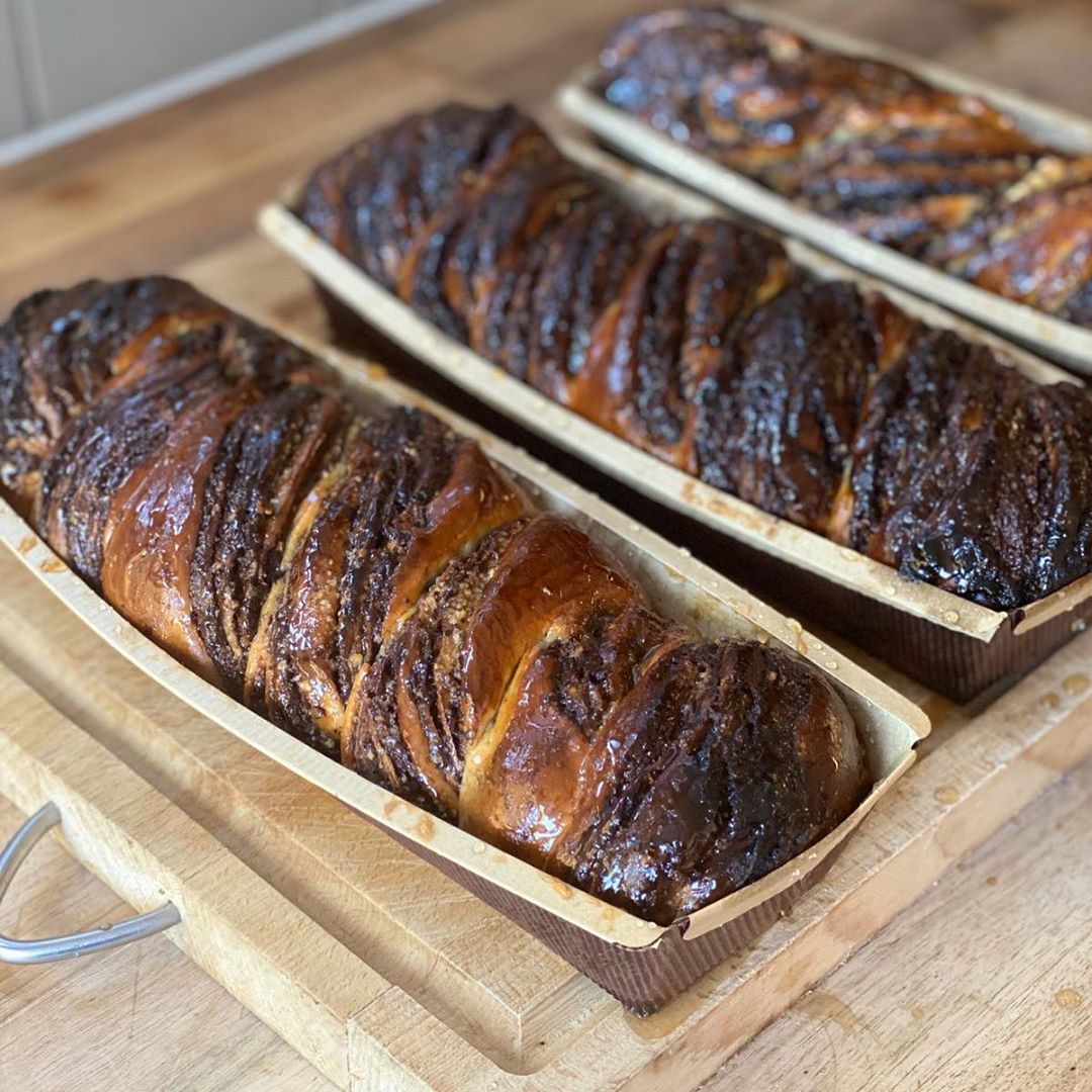 Three golden-brown braided challah loaves with glossy, caramelized crusts cooling in wooden baking molds on a kitchen counter.