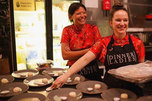 Two smiling women in red traditional Chinese tops prepare dumplings in a kitchen, with rows of uncooked dumplings on dark plates.
