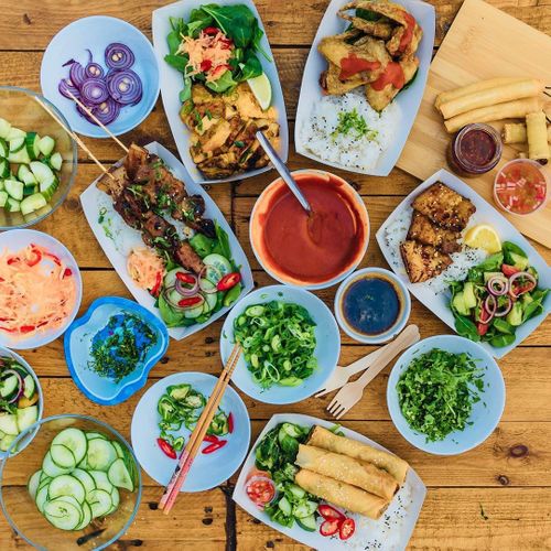 A wooden table filled with colorful Filipino dishes including grilled meats, fresh vegetables, rice, spring rolls, and various dipping sauces served on blue and white plates.