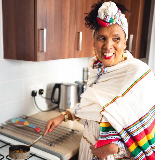 A smiling woman in traditional Ethiopian white dress with colorful trim and headwrap stands in a modern kitchen.