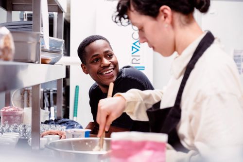 Two chefs working together in a professional kitchen, with one chef smiling while preparing food at a stainless steel counter.
