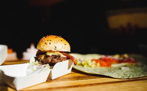 A gourmet burger with sesame seed bun and bacon sits on parchment paper on a wooden cutting board alongside fresh salad.