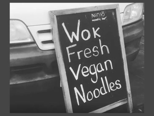 A black chalkboard sign advertising "Wok Fresh Vegan Noodles" sits on a street outside Ninjō Noodle Bar.
