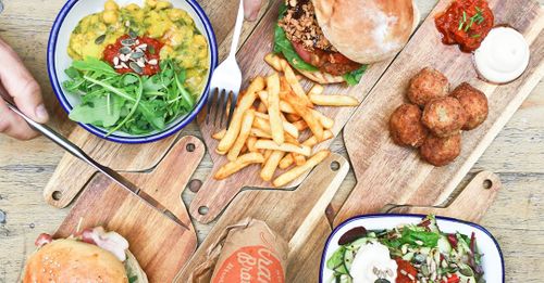 A spread of Italian dishes on wooden boards including arancini balls, burgers, pasta salad, and fries arranged on a rustic table.