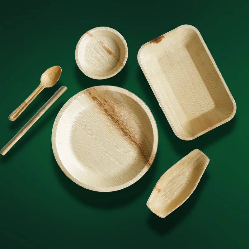 Biodegradable palm leaf plates, bowls, and wooden utensils arranged on a green background showcasing eco-friendly packaging.