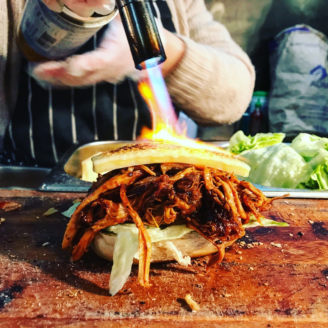 A chef uses a kitchen torch to caramelize a gourmet burger topped with crispy fried onions on a wooden cutting board.