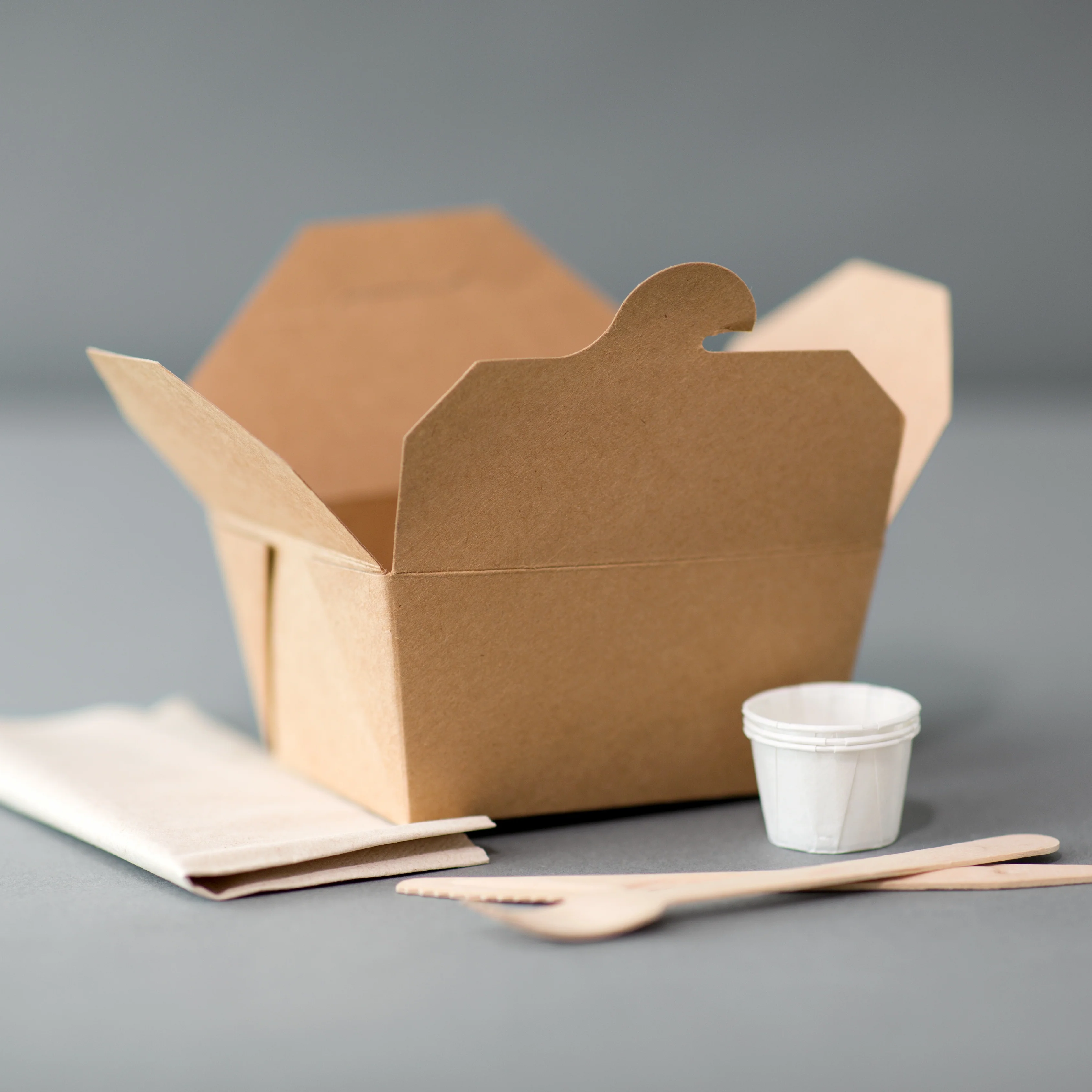 Brown takeout containers with wooden utensils and sauce cup arranged on a gray surface.