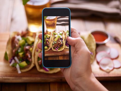 Hand holding smartphone taking photo of street tacos on wooden board with beer and salsa in background.