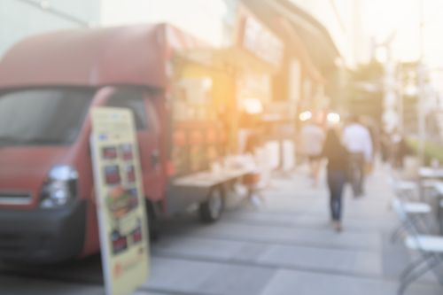 A red food truck parked on a sunny city street with a menu board displayed outside and pedestrians walking by.