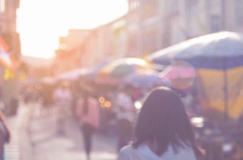 Blurred scene of people walking through a bustling outdoor market with colorful umbrellas and sunlight streaming through.