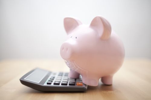 Pink piggy bank sitting on top of a black calculator on a wooden surface against a white background.